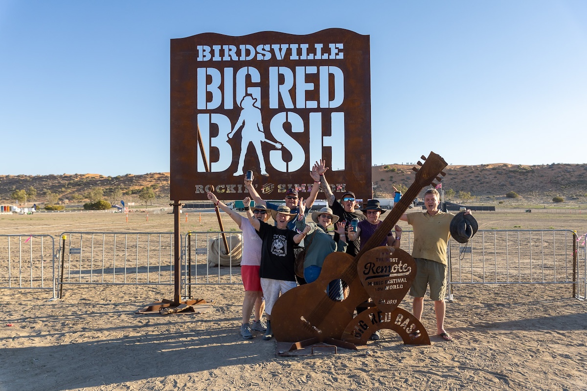 Happy festival-goers posing beneath the Birdsville Big Red Bash welcome sign in the desert outback.